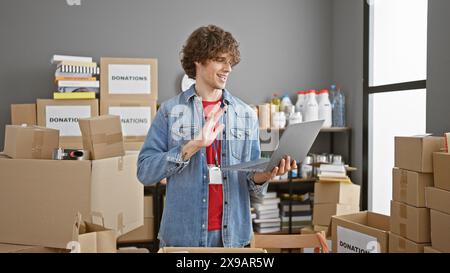 Un giovane amichevole con i capelli ricci si trova in mezzo a scatole di cartone in un magazzino, utilizzando un computer portatile e gestando. Foto Stock