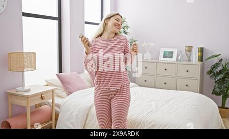 Una giovane donna gioiosa in pigiama a righe danzano in una camera da letto accogliente ed elegante con una parete viola chiaro. Foto Stock