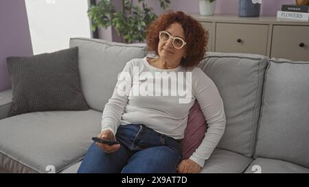Un'anziana donna caucasica con capelli ricci e occhiali si siede comodamente su un divano del soggiorno reggendo un telecomando tv, in un accogliente ambiente domestico interno. Foto Stock