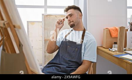 Uomo con barba in grembiule di denim che contempla in uno studio d'arte con tele e oggetti d'arte intorno. Foto Stock