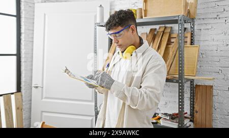 Un giovane ispeziona un appunti in un'officina ben organizzata per la lavorazione del legno indossando indumenti protettivi. Foto Stock