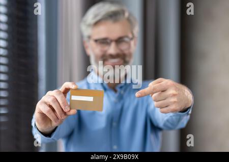 Uomo di mezza età con barba e bicchieri sorridente, che tiene e indica una carta di credito, in casa Foto Stock