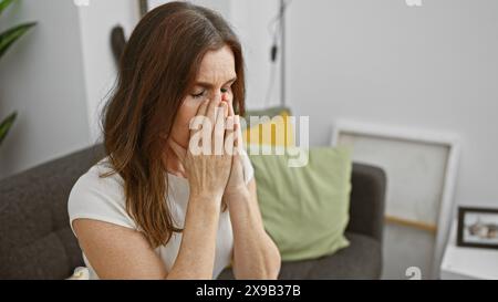 Donna matura in difficoltà con le mani che coprono il viso seduta sul divano a casa, trasmettendo emozioni di tristezza o preoccupazione. Foto Stock