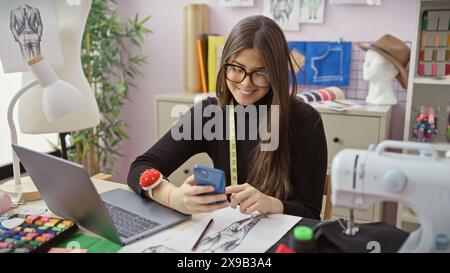 Donna sorridente che usa lo smartphone in un moderno negozio di sartoria con macchina da cucire e schizzi alla moda Foto Stock