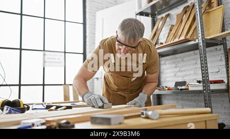 Un uomo maturo dai capelli grigi lavora con attenzione in un laboratorio di falegnameria circondato da legno e utensili. Foto Stock