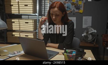 Una donna rossa concentrata lavora diligentemente nell'ufficio di un detective disordinato, circondato da prove di casi ed effetti personali. Foto Stock