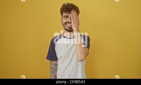 Sorridente giovane ispanico con barba e tatuaggi in piedi su uno sfondo giallo, toccando giocosamente il suo viso. Foto Stock