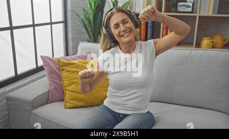 Una donna gioiosa di mezza età che indossa le cuffie balla su un divano in un salotto accogliente ascoltando musica. Foto Stock