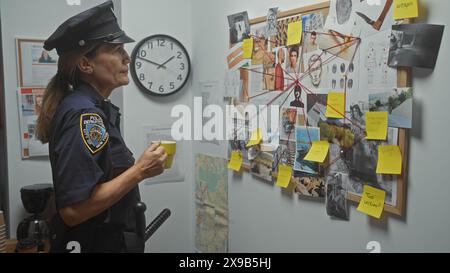 Un agente di polizia di mezza età che tiene una tazza di caffè analizzando un muro di prove in una stanza simile a un ufficio. Foto Stock