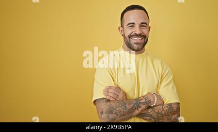 Bellissimo giovane ispanico con barba e tatuaggi che si posa con sicurezza su un vibrante sfondo giallo. Foto Stock