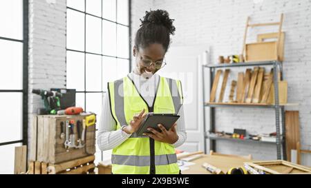 Donna afroamericana sorridente che usa un tablet in un laboratorio di falegnameria, ritrae l'abilità artigianale e l'integrazione tecnologica. Foto Stock