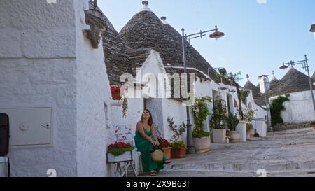 Una bella giovane donna ispanica con un abito verde siede e sorride su una strada affascinante di alberobello, in italia, circondata da tradizionali case di trulli Foto Stock