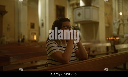 Una giovane donna ispanica è vista pregare all'interno di una bella chiesa italiana, fornendo un ambiente religioso sereno. Foto Stock