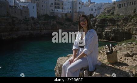 Giovane donna ispanica sorridente su una scogliera di roccia a polignano a mare, puglia, italia, seduta sul mare con un tradizionale ambiente da spiaggia sullo sfondo. Foto Stock