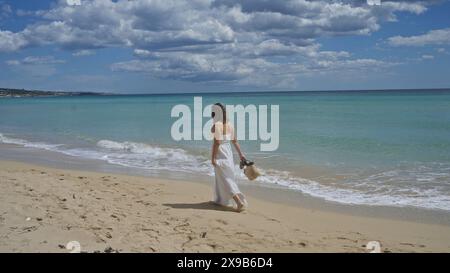 Una giovane e bella donna ispanica passeggia lungo la tranquilla spiaggia di pescoluse, salento, puglia, italia, godendosi le dolci onde e le pittoresche terre Foto Stock