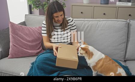 Una donna sorridente si siede su un divano a casa, disimballando una scatola con il suo attento chihuahua che guarda. Foto Stock