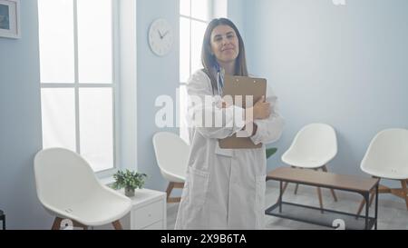 Una giovane e attraente dottoressa con i capelli bruna si trova in una sala d'attesa della clinica, tenendo in mano un appunti, indossando un camice bianco da laboratorio, al chiuso Foto Stock