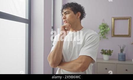 Un giovane uomo ispanico contemplativo sorge con cura in una camera moderna vestita con abbigliamento casual bianco. Foto Stock