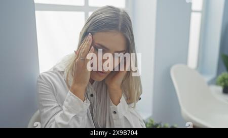 Una giovane donna bionda caucasica con una camicia bianca tiene la testa all'interno di una clinica, riflettendo lo stress o il mal di testa. Foto Stock