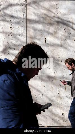 Due uomini che camminano all'aperto, entrambi invischiati nei loro smartphone, proiettano ombre su un muro strutturato dietro di loro. Foto Stock