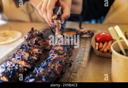 Un primo piano di costolette di maiale grigliate viene raccolto per mano in un popolare ristorante, con un lato di salsa e verdure arrostite, mettendo in risalto l'esperienza culinaria saporita e rustica. Nonostante la guerra, gli attacchi missilistici e le incursioni aeree, la vita pacifica continua nella città Ucraina di Leopoli. Nonostante le difficoltà e i pericoli, i residenti della città stanno cercando di mantenere un ritmo di vita normale, lavorando, socializzando, rilassandosi e prendendosi cura l'uno dell'altro. Questo dimostra la forza dello spirito e la determinazione del popolo a mantenere una vita normale in condizioni di guerra e aggressione. Foto Stock