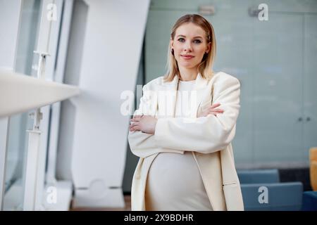 Bella donna d'affari bionda incinta con un abbigliamento elegante e alla moda un abito bianco e un blazer che guarda la fotocamera in un'azienda Foto Stock