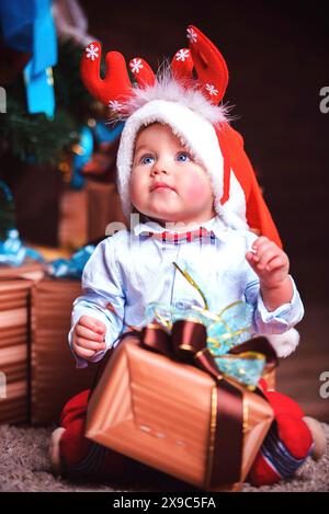 Un bambino vestito in abiti festivi seduto vicino a un albero di Natale con in mano una confezione regalo Foto Stock