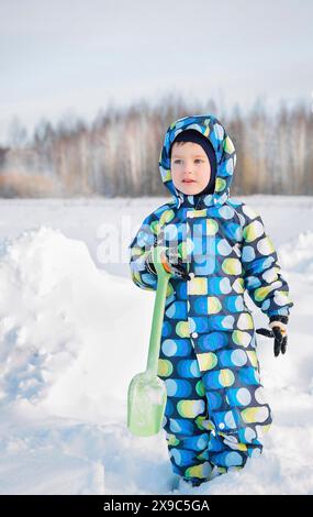 Bambino in un vestito colorato in piedi sulla neve tenendo in mano una pala verde in una giornata invernale con alberi sullo sfondo Foto Stock