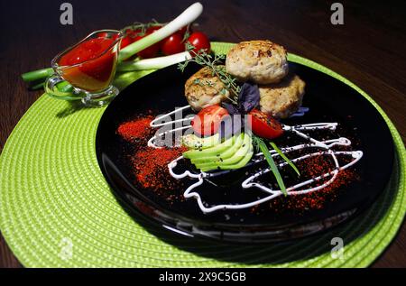 Tre polpette su un piatto nero guarnite con avocado, pomodori ciliegini, erbe aromatiche, decorazioni artistiche in salsa, accompagnate da un piccolo piatto di immersione Foto Stock