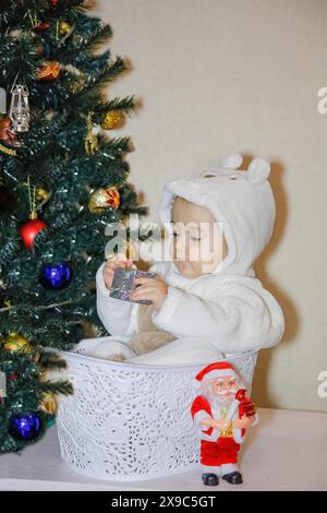 Un bambino in abiti bianchi si siede in un cestino bianco accanto a un albero di Natale decorato e a una statuetta di Babbo Natale Foto Stock