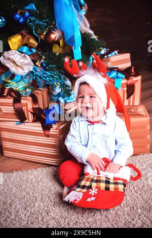 Un bambino gioioso che indossa un cappello di Babbo Natale e si siede davanti a un albero di Natale circondato da regali Foto Stock