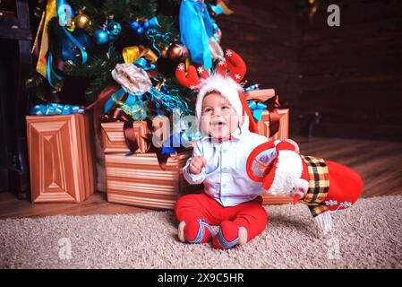 Un bambino in un cappello di Babbo Natale e corna di renna si siede gioiosamente accanto a un albero di Natale e regali Foto Stock