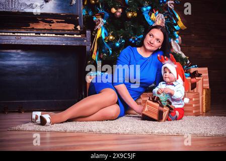 Una donna in abito blu siede accanto a un albero di Natale con un bambino in un cappello di Babbo Natale, circondato da regali Foto Stock