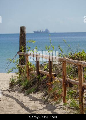 Vista di una spiaggia sabbiosa con ringhiera in legno in primo piano e una nave all'orizzonte, sgorga sulla costa polacca del Mar Baltico con alberi verdi e. Foto Stock