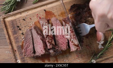 Lo chef affetta carne di manzo succosa grigliata con un coltello su un tagliere di legno in cucina, primo piano Foto Stock