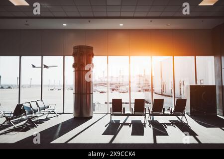 Interno di un terminal aeroportuale con aree salotto vuote, grandi finestre di vetro che si affacciano sulla pista e un aereo che decolla sullo sfondo. TH Foto Stock