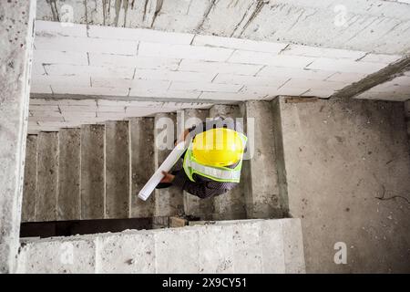 giovane ingegnere professionista in casco protettivo e blueprints carta a portata di mano presso il cantiere di costruzione della casa Foto Stock