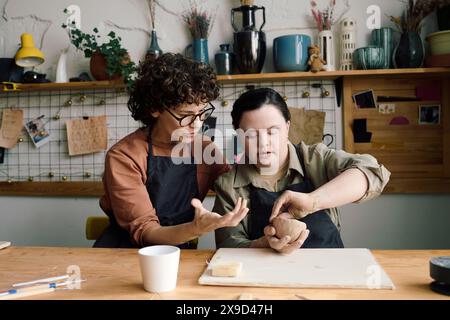 Donna matura che indossa gli occhiali seduti a tavola insegnando al suo allievo con la sindrome di Down come modellare l'argilla Foto Stock