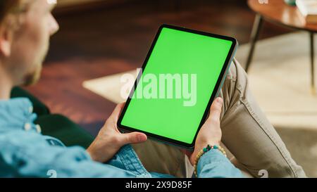 Giovane uomo che tiene in mano un tablet con schermo di mascheratura con schermo verde. Uomini che si rilassano a casa, guardano video e leggono post sui social media su dispositivi mobili. Primo piano con riprese statiche sulla spalla. Foto Stock