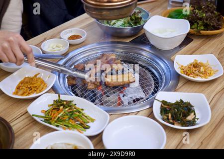 Ulsan, Corea del Sud - 24 febbraio 2024: Una persona che usa le pinze per affumicare la carne su una griglia a carbone, con il fumo che viene aspirato in una presa d'aria sopra, sur Foto Stock