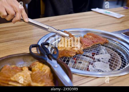 Ulsan, Corea del Sud - 24 febbraio 2024: Una mano che usa le pinze per accendere la carne su una griglia a carbone, con carne cruda pronta per essere cucinata in primo piano in una Foto Stock