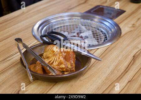 Ulsan, Corea del Sud - 24 febbraio 2024: Un piatto di carne pronta da cucinare con forbici e pinze accanto a una griglia a carbone su un tavolo in un samgyeopsal re Foto Stock