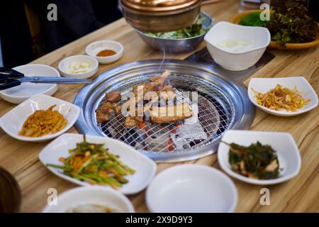 Ulsan, Corea del Sud - 24 febbraio 2024: Fumo che sale dalla carne alla griglia su una griglia a carbone e viene aspirato in una bocca sopra, circondato da banchan Foto Stock