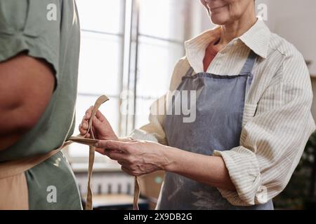 Vista laterale ravvicinata di due donne mature che legano grembiuli in uno studio d'arte in preparazione per la lezione di ceramica Foto Stock