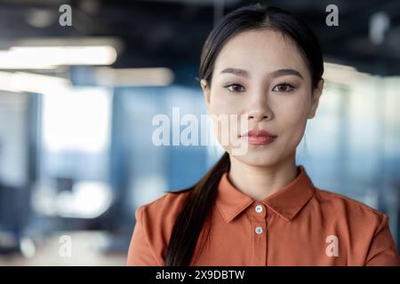 Primo piano ritratto di una donna asiatica seria e sicura di sé sul posto di lavoro all'interno dell'ufficio che guarda con attenzione alla fotocamera, mentre una donna lavora in ufficio si avvicina alla finestra. Foto Stock