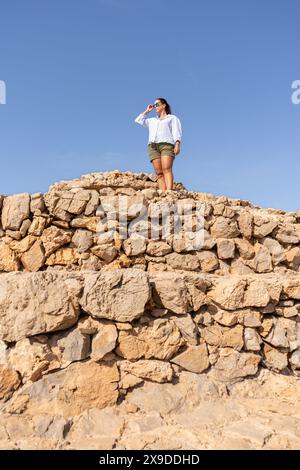 Una giovane donna con occhiali da sole e abbigliamento casual si erge con sicurezza su antiche pareti di pietra contro un cielo azzurro, catturando l'essenza dell'avventura A. Foto Stock