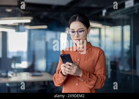 Donna asiatica che usa il telefono, donna d'affari seria e premurosa all'interno dell'ufficio che digita messaggi di testo e naviga sui social media, sviluppatrice di software donna che prova app online. Foto Stock