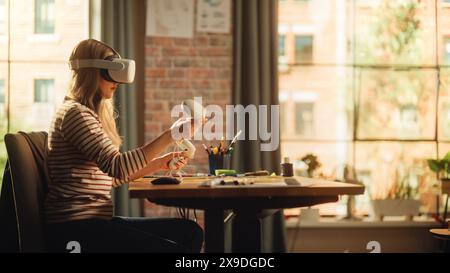 Donna bionda che indossa le cuffie VR 3D e utilizza i moderni controller nel soggiorno dell'appartamento Loft. Designer donna che lavora in applicazioni futuristiche. Nuove tecnologie e concetto di realtà aumentata. Foto Stock