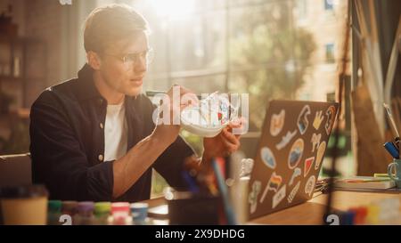 Talentuoso artista maschile che lavora su Colored Design for Sneakers, utilizza un computer portatile in Loft Office Studio. Coppia di scarpe personalizzata da stilista di moda. Concetto di post-produzione. Foto Stock