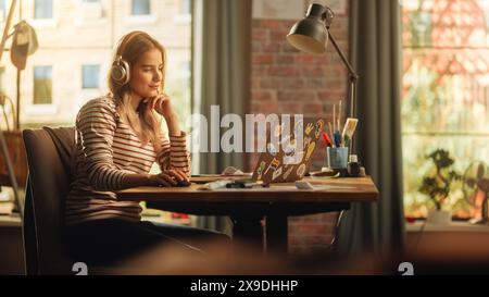Giovane e bella donna con i capelli biondi che indossa le cuffie e lavora da casa su un computer portatile nell'elegante appartamento Loft. Creative Female sorridente, controllare i social media, navigare in Internet. Foto Stock
